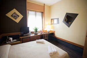 A hotel room with a bed, a desk with a television, a chair, a lamp, and a window with curtains.