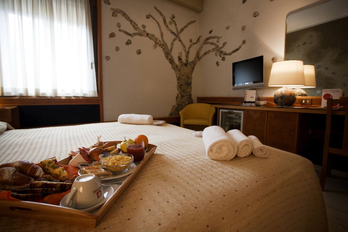 A hotel room with a bed, a desk, a chair, a TV, and a tray with breakfast items.