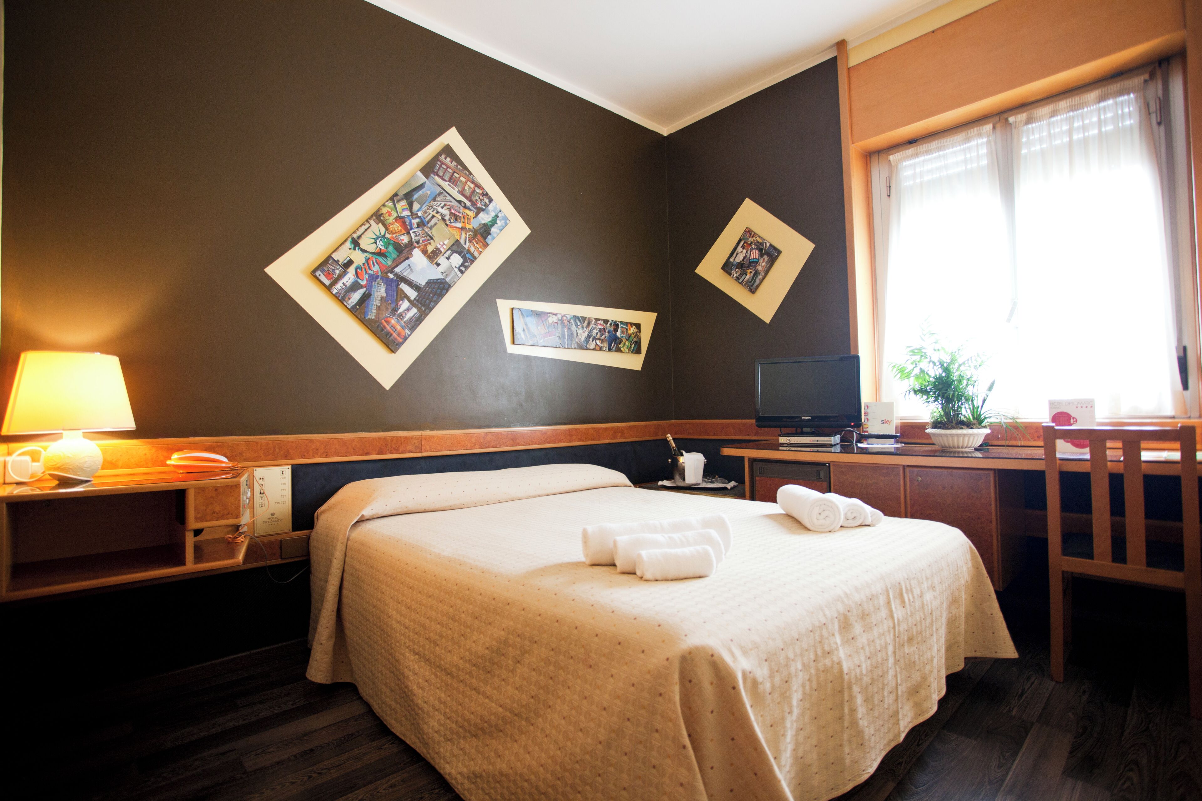 A hotel room with a large bed, a desk with a computer, a chair, a lamp, and two framed pictures on the wall.