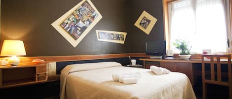 Ein Hotelzimmer mit einem großen Bett, einem Schreibtisch mit Computer, einem Sessel, einer Lampe und zwei gerahmten Bildern an der Wand.