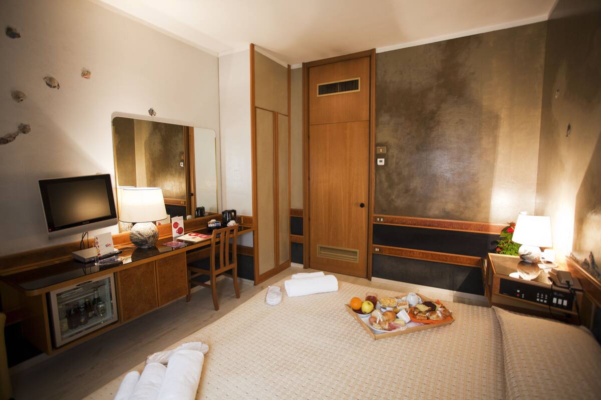 A hotel room with a bed, a desk with a computer, a television, a minibar, and a bathroom with a shower and toilet.