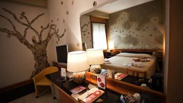 Ein Hotelzimmer mit einem Bett, zwei Lampen, einem Schreibtisch mit Stuhl, einem Spiegel und einer Wand mit dekorativem Grün.