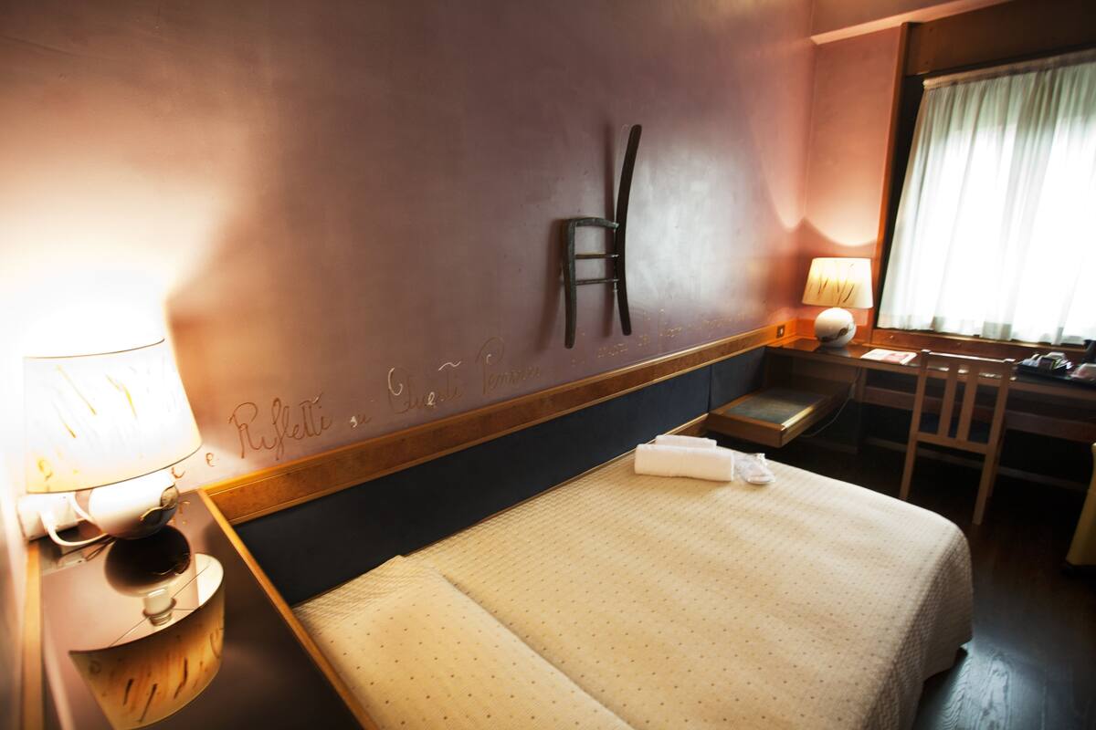 A hotel room with a bed, a desk, a chair, a lamp, and a window with curtains.