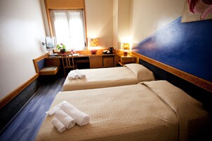 Hotellihuone, jossa on kaksi sänkyä, työpöytä, tuoli, pieni pöytä ja ikkuna, jossa on verhot.