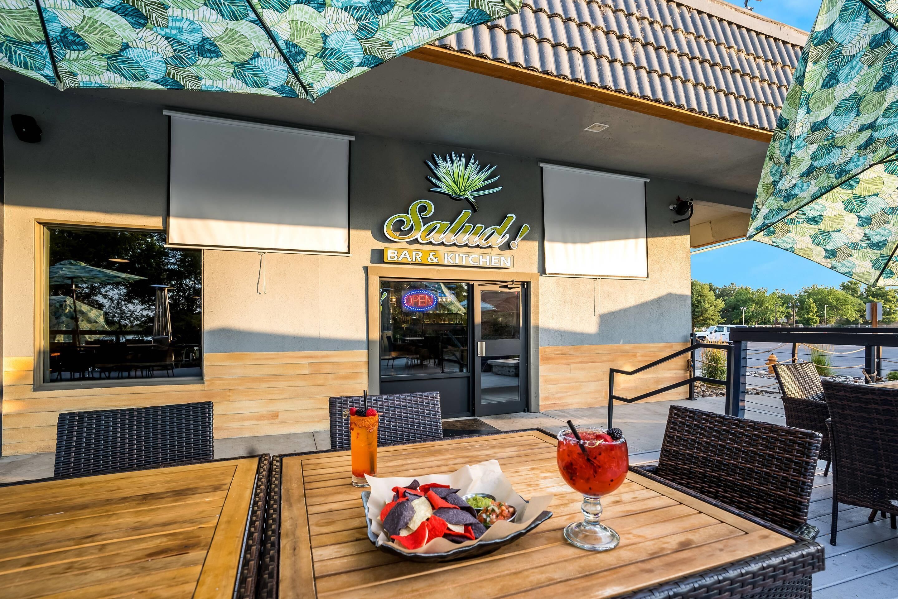 Un área de estar al aire libre con una mesa para dos, un menú y un edificio con un cartel que dice 'Salud! BAR & KITCHEN'.