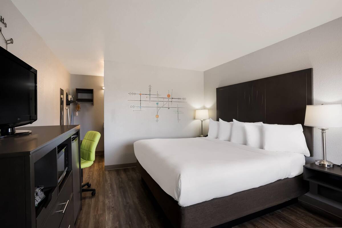 Ein Hotelzimmer mit Bett, Schreibtisch mit Fernseher, Stuhl, Wandbild und Nachttisch mit Lampe.