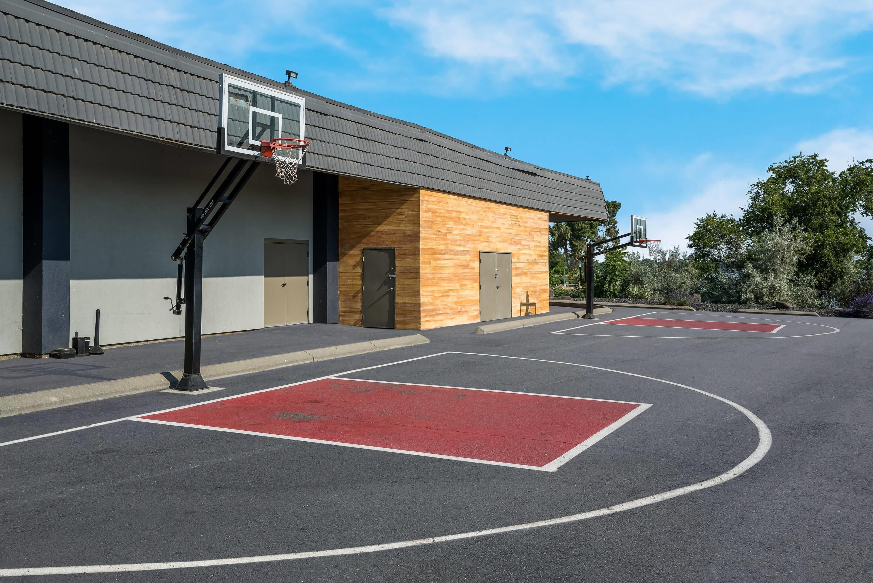 An outdoor basketball court with two hoops, a building with a wooden facade, and a clear sky.