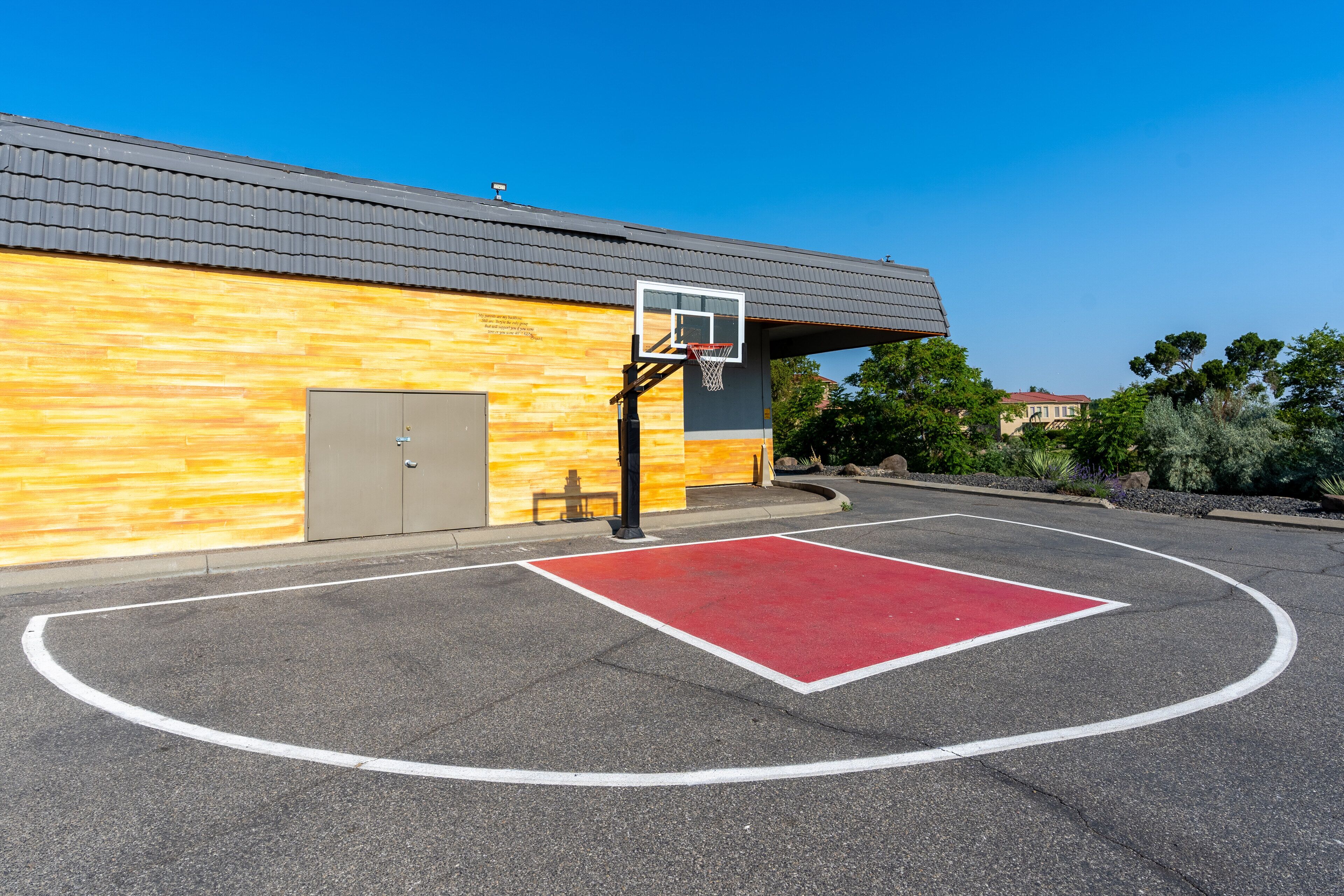 An outdoor basketball court with a red surface, a basketball hoop, and a building with a wooden facade in the background.