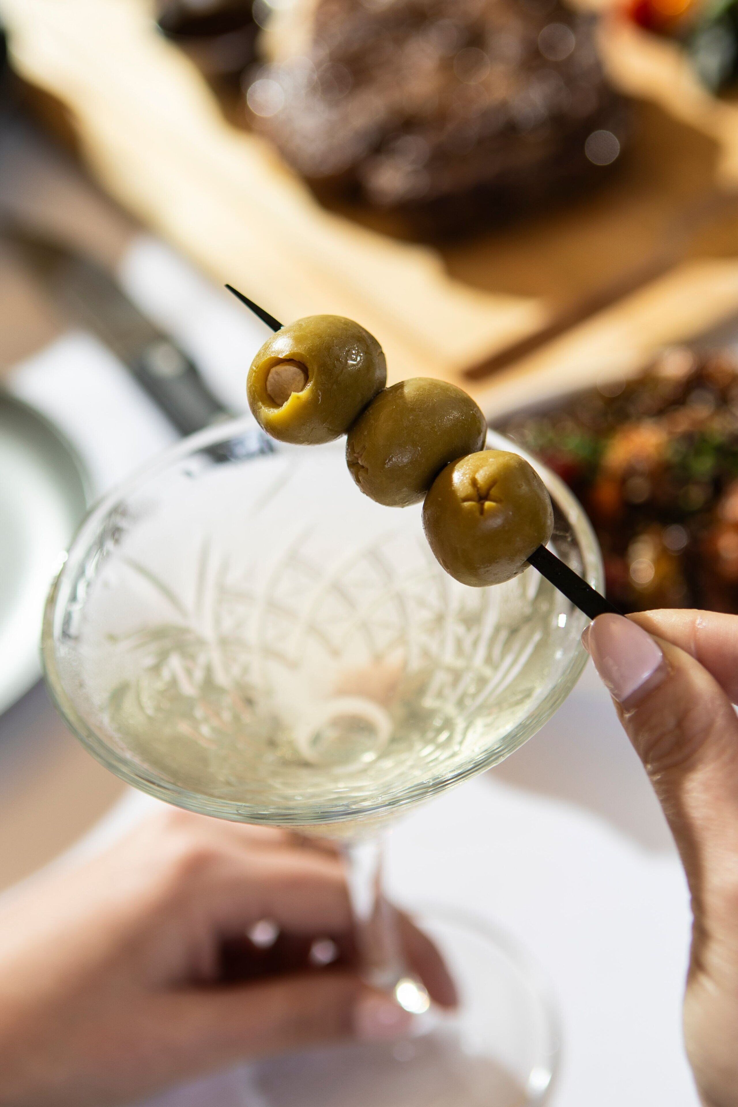 A hand holding a glass with three olives skewered on a stick.
