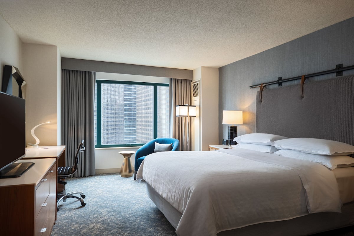 A hotel room with a large bed, a desk with a computer, a chair, a lamp, and a view of a cityscape through a window.