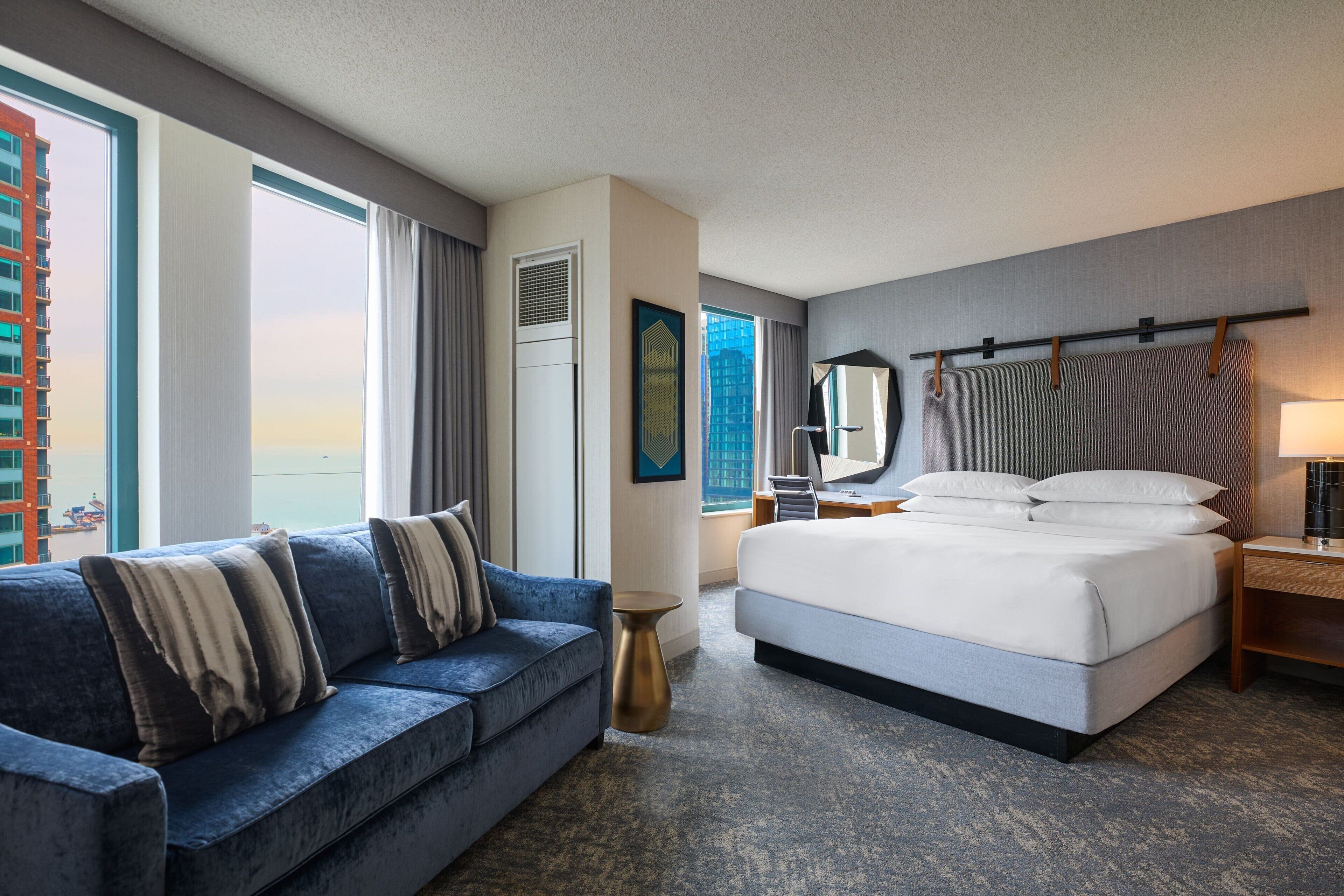 Camera d'hotel con un letto grande, una poltrona, un tavolino e uno specchio, con vista su un edificio.