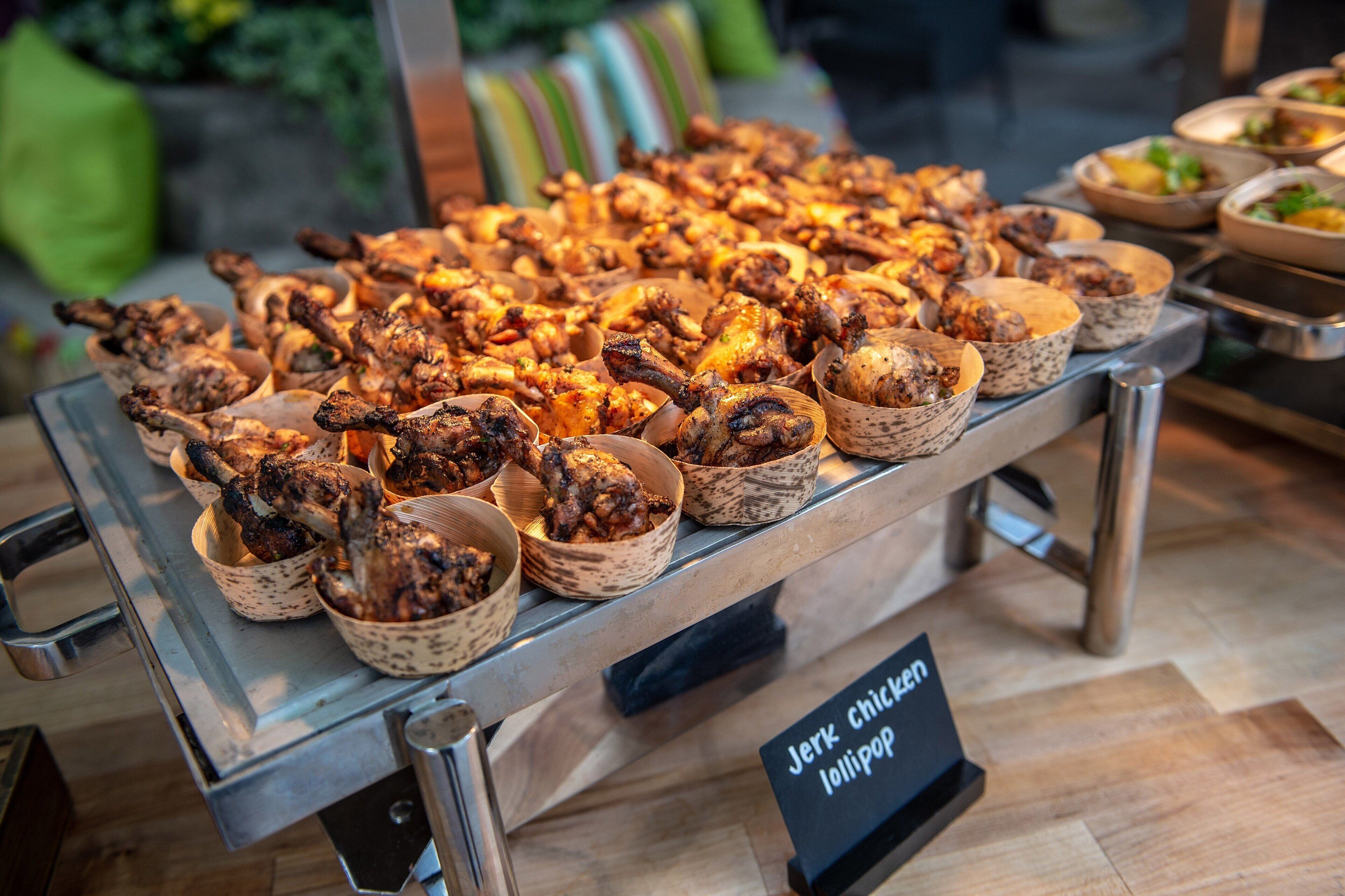 Une table de buffet proposant divers plats, notamment du poulet à la sauce jerk et des brochettes.