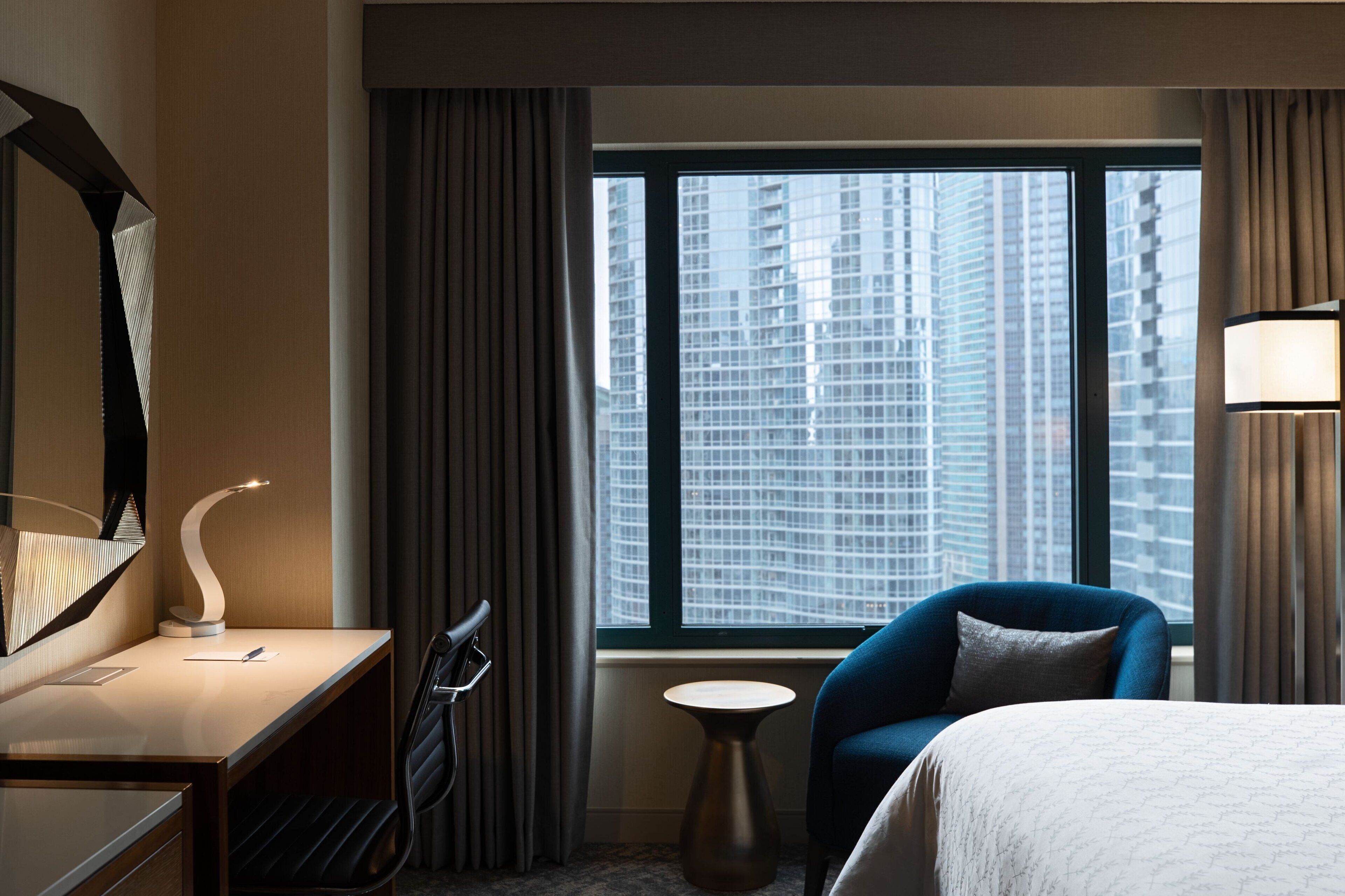 A hotel room with a bed, a desk, a chair, a small table, and a view of tall buildings through a large window.