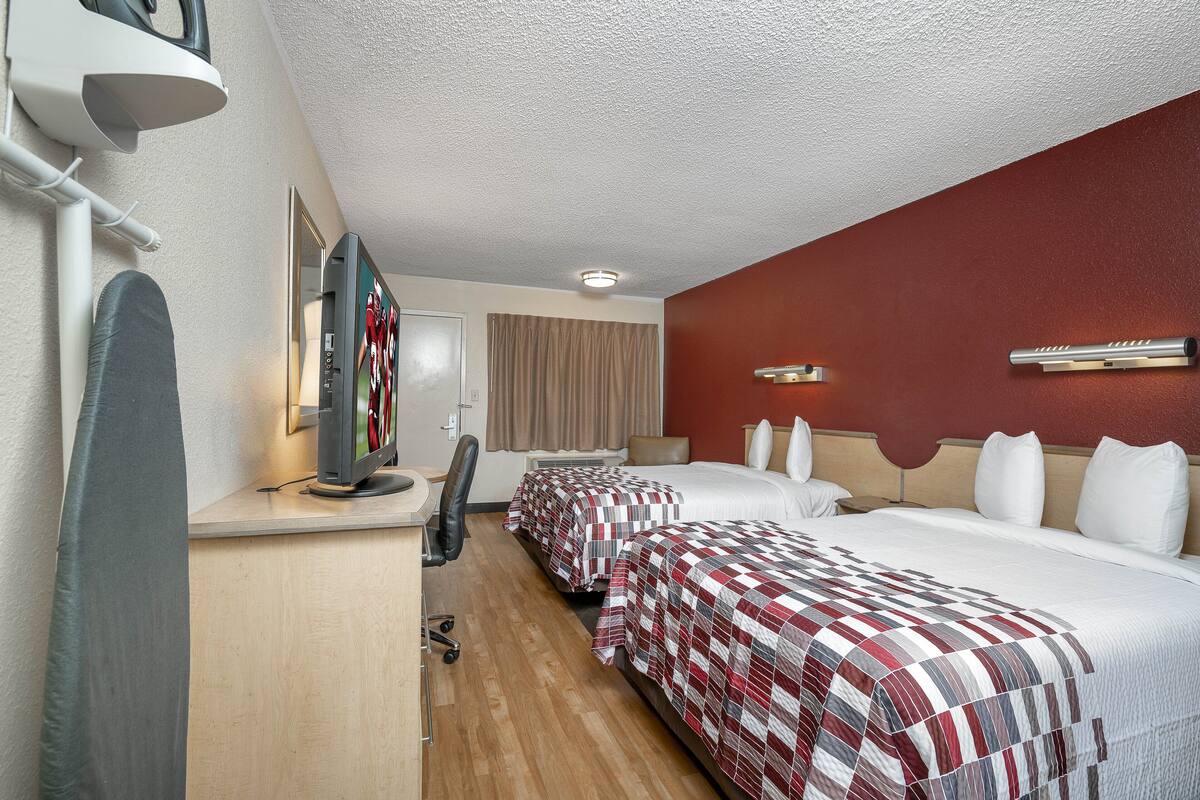 A hotel room with two beds, a desk with a computer, a chair, a TV, and a window with curtains.