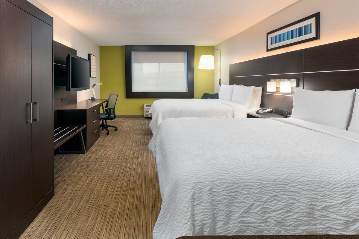 A hotel room with two beds, a desk with a chair, a television, and a large window with blinds.