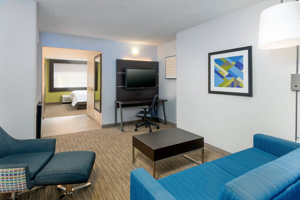 A hotel room with a TV, a desk, two blue sofas, a small table, and a bed with a window.