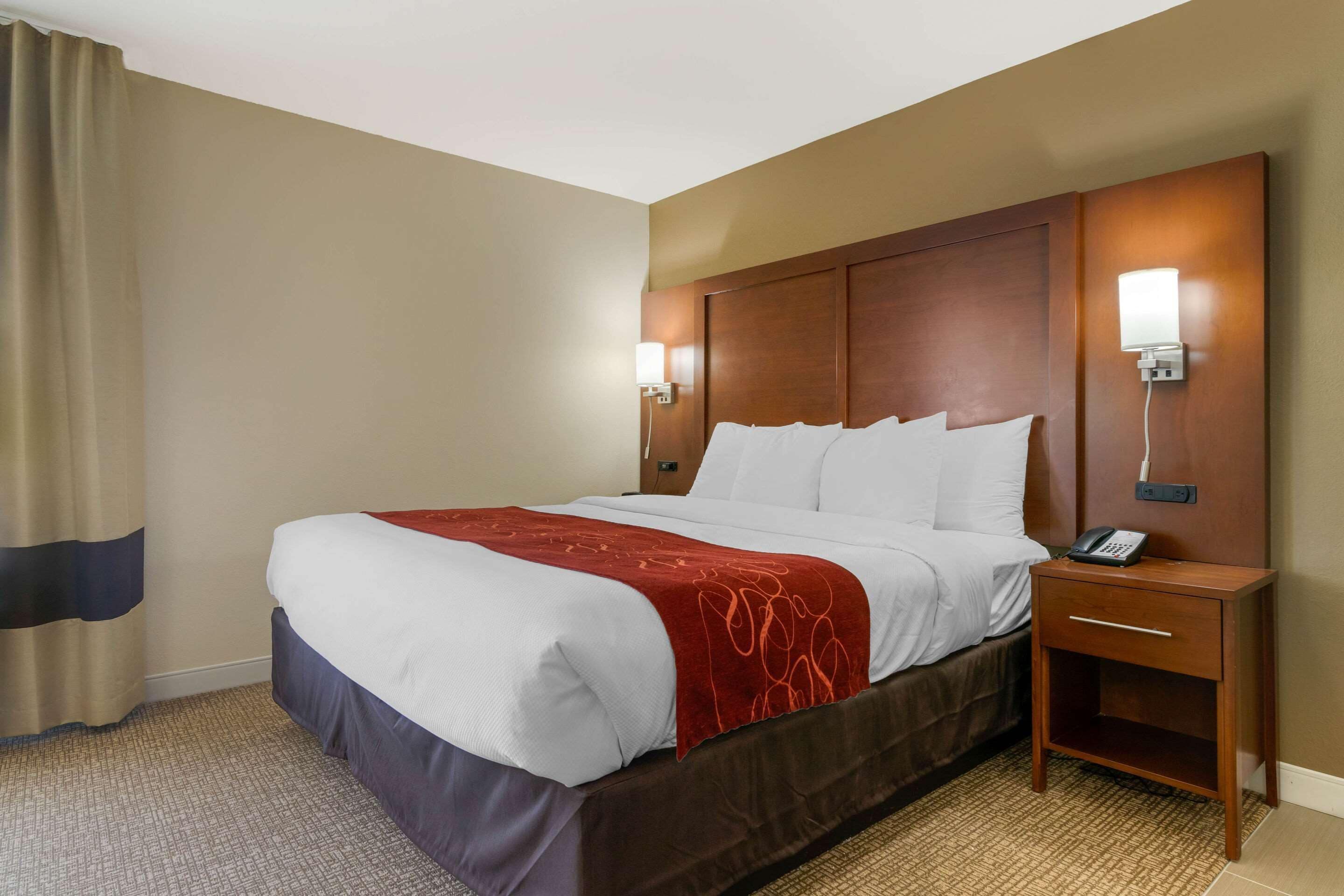A hotel room with a large bed, a wooden nightstand with a phone, and wall-mounted lights.