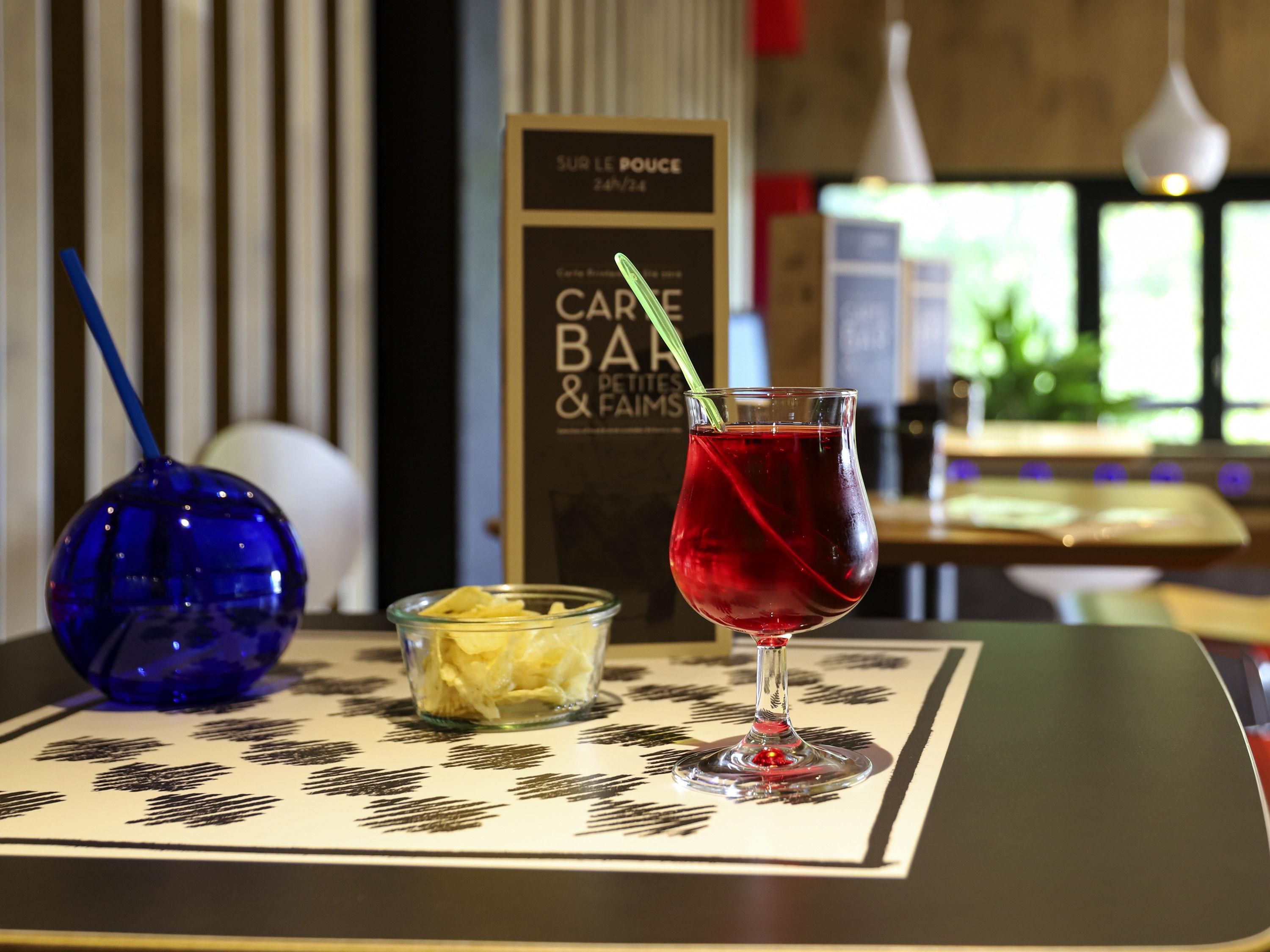 Een tafel met een glas rode drank, een blauwe vaas en een schaaltje chips.