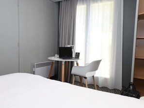 Habitación de hotel con cama, escritorio con una computadora portátil, silla, maleta y ventana con cortinas.