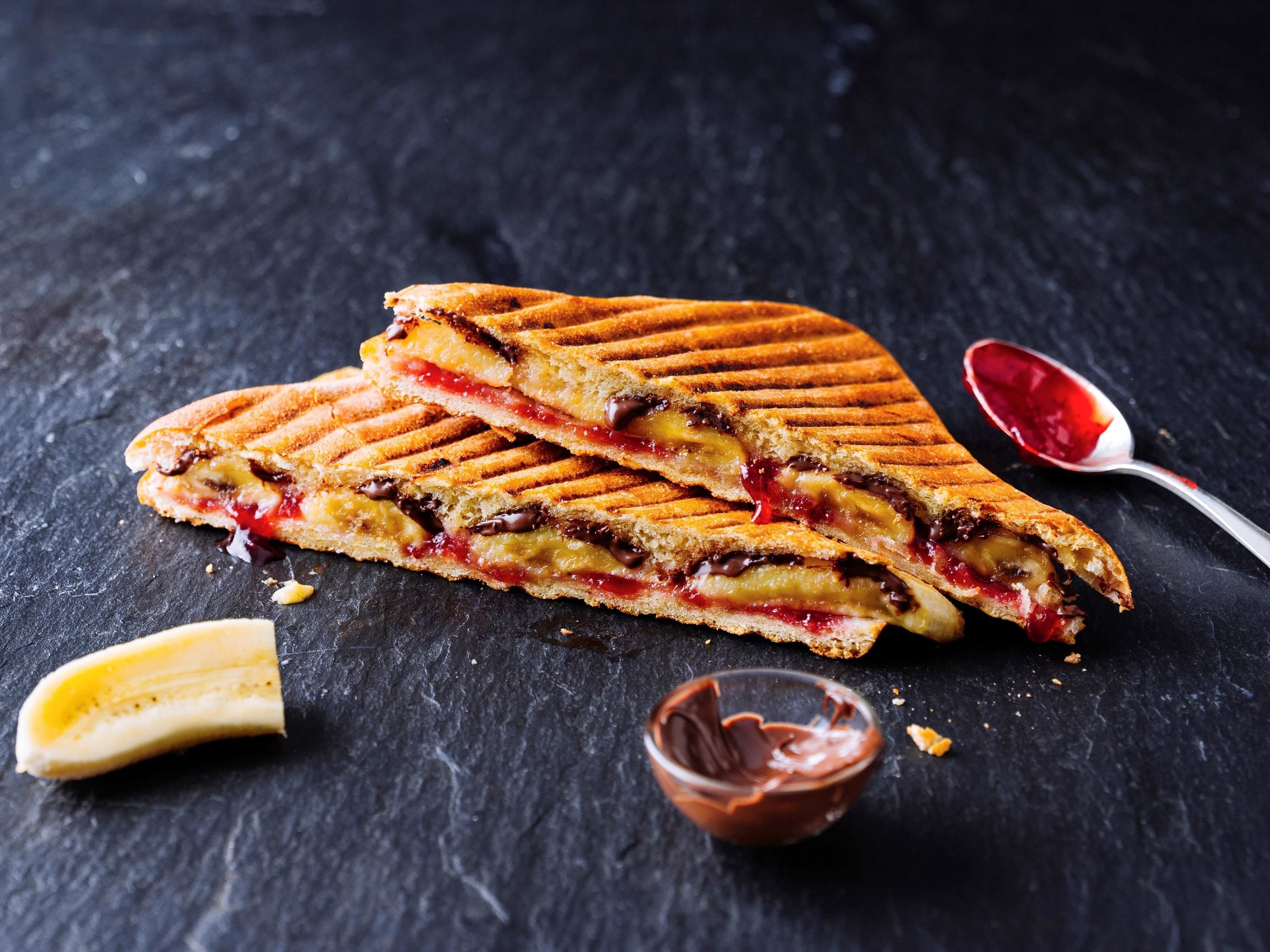 Ein gegrilltes Sandwich mit sichtbaren Zutaten, ein Löffel Marmelade und eine kleine Schale Schokoladenaufstrich auf einer dunklen Oberfläche.