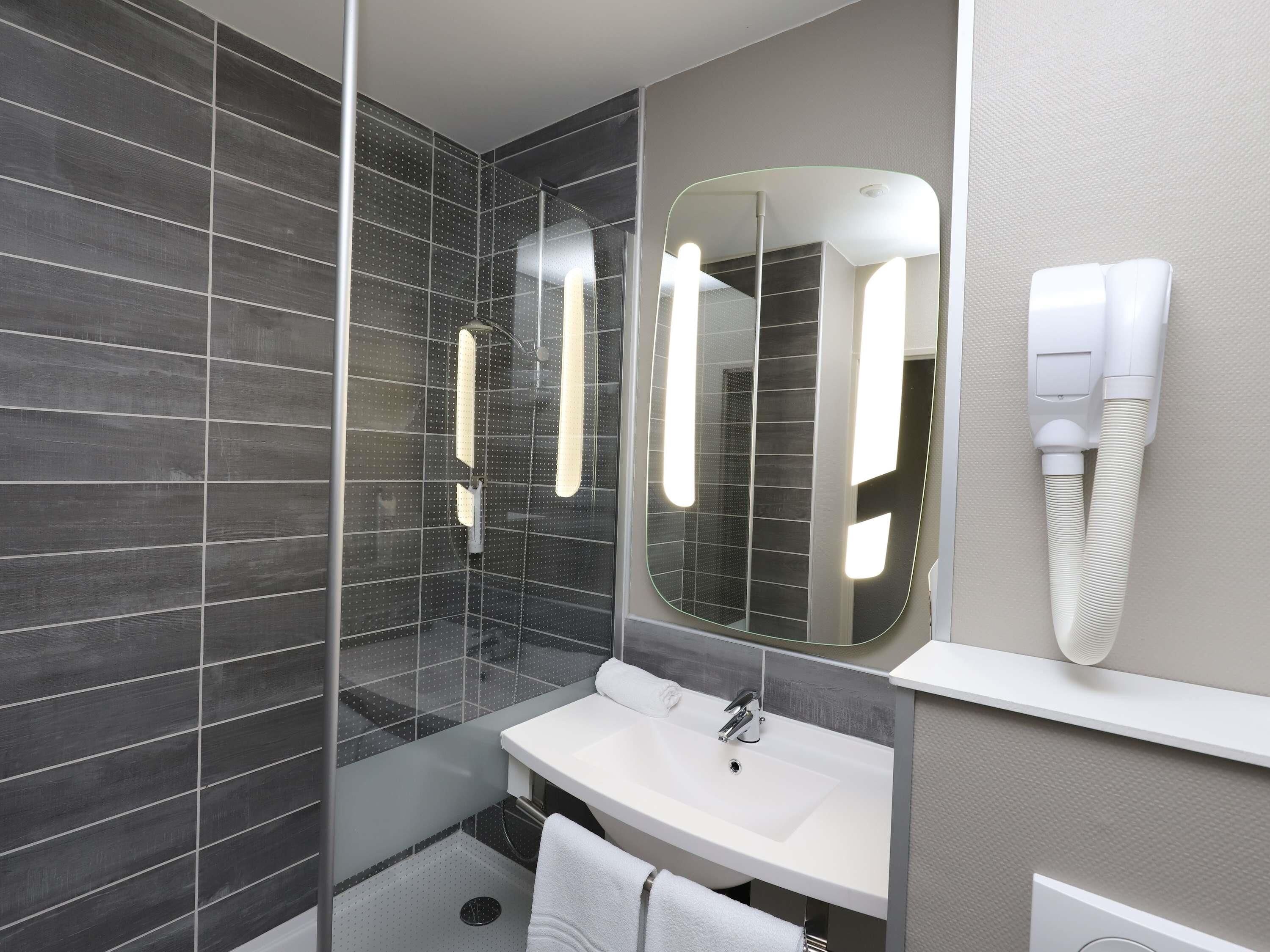 A modern bathroom with a glass shower enclosure, a white sink, a large mirror, and a hairdryer mounted on the wall.