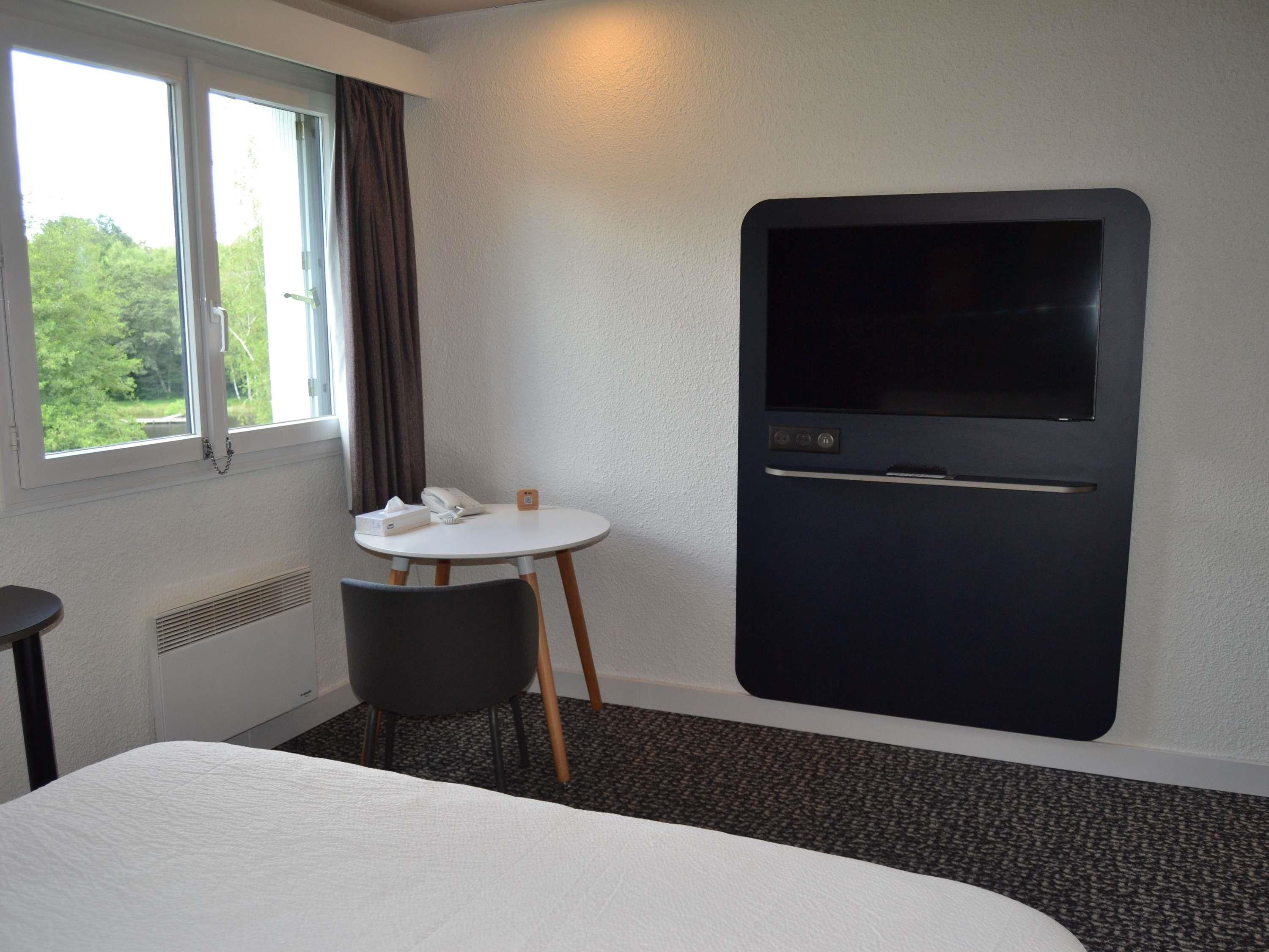 A hotel room with a bed, a small round table with a telephone and tissues, a TV mounted on the wall, and a window with curtains.