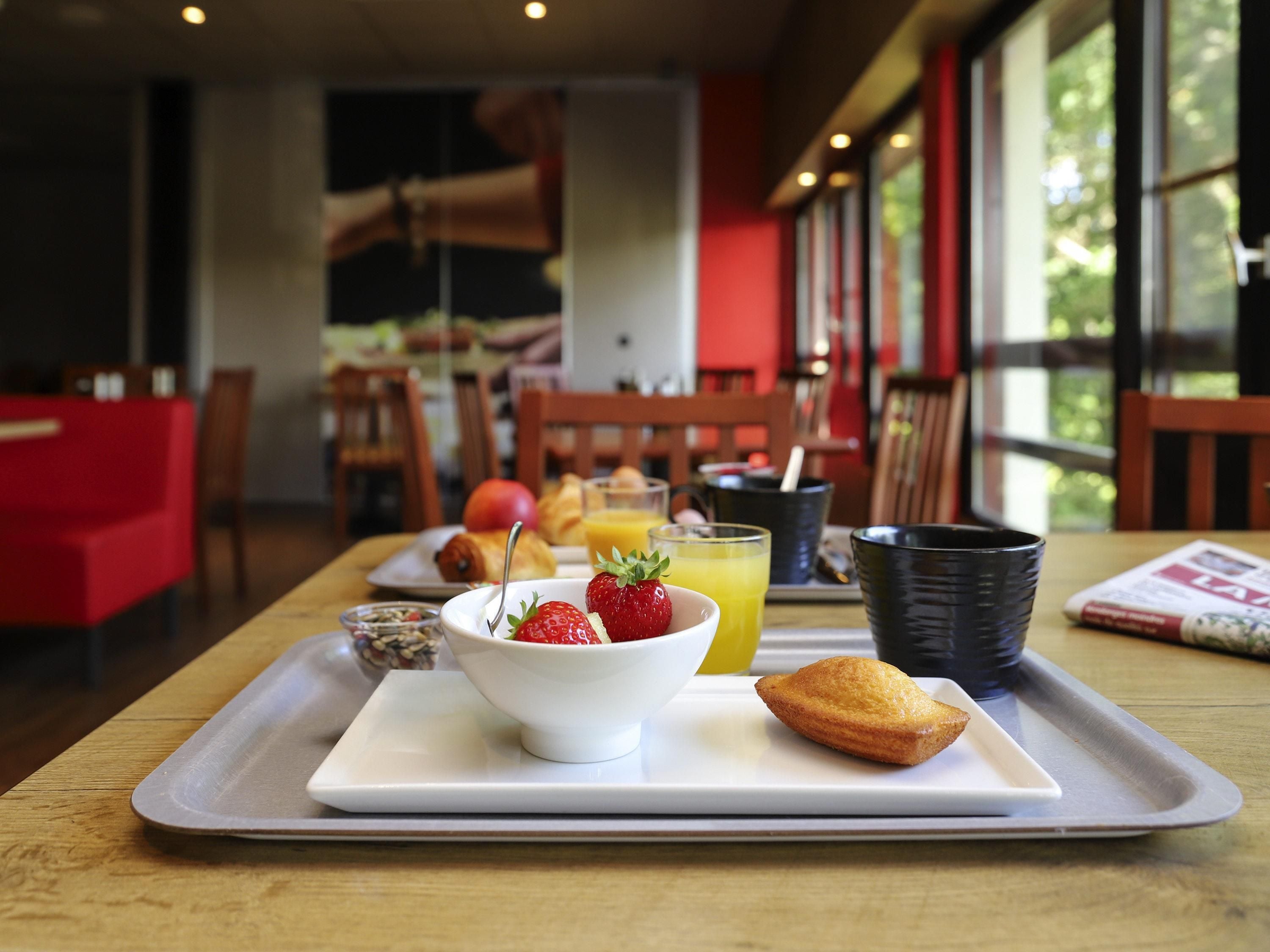 A breakfast tray with a croissant, strawberries, and orange juice on a wooden table in a restaurant with red chairs and large windows.