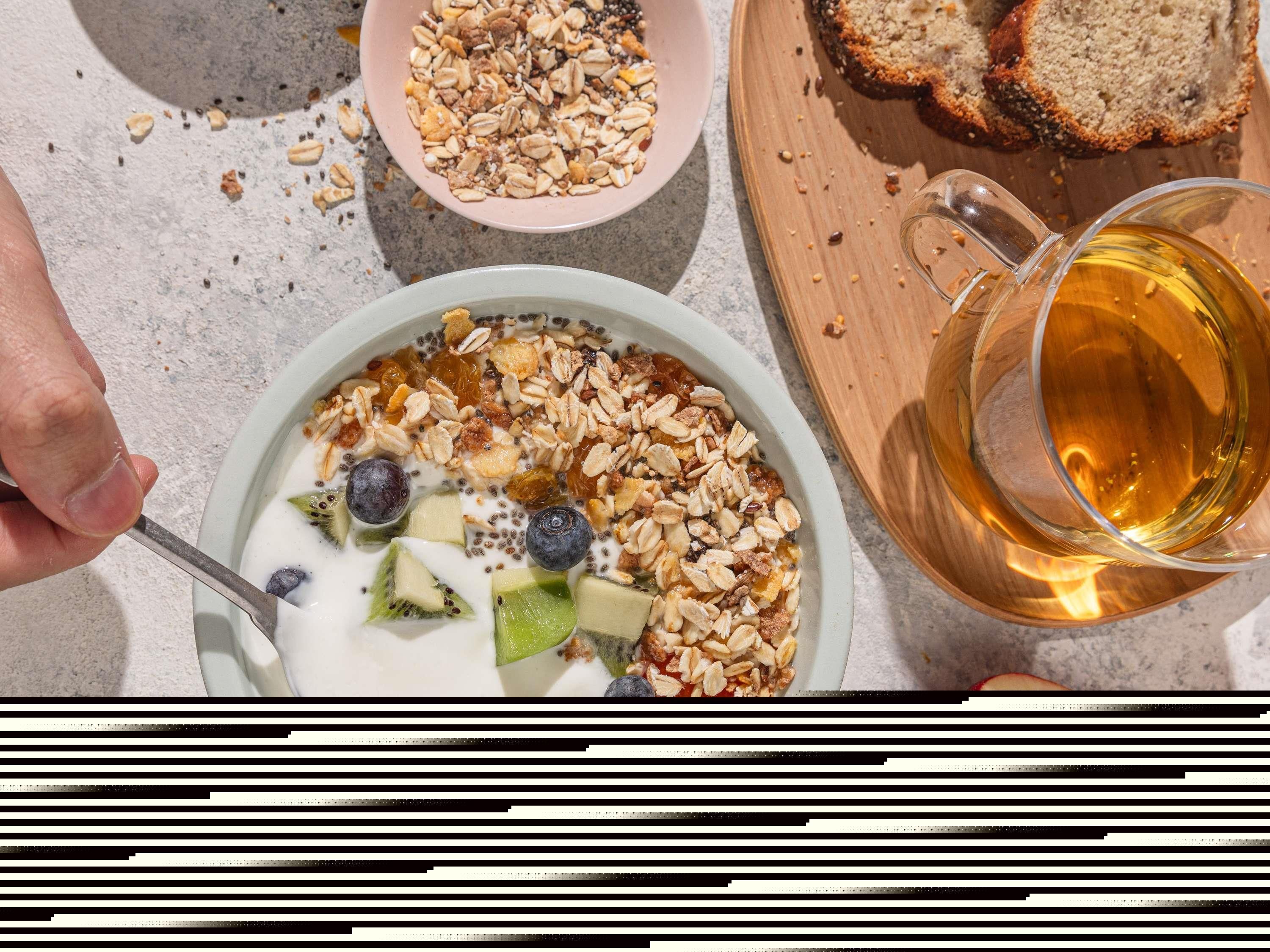 A bowl of yogurt with granola and fruit, a glass of amber liquid, and slices of bread on a wooden board.