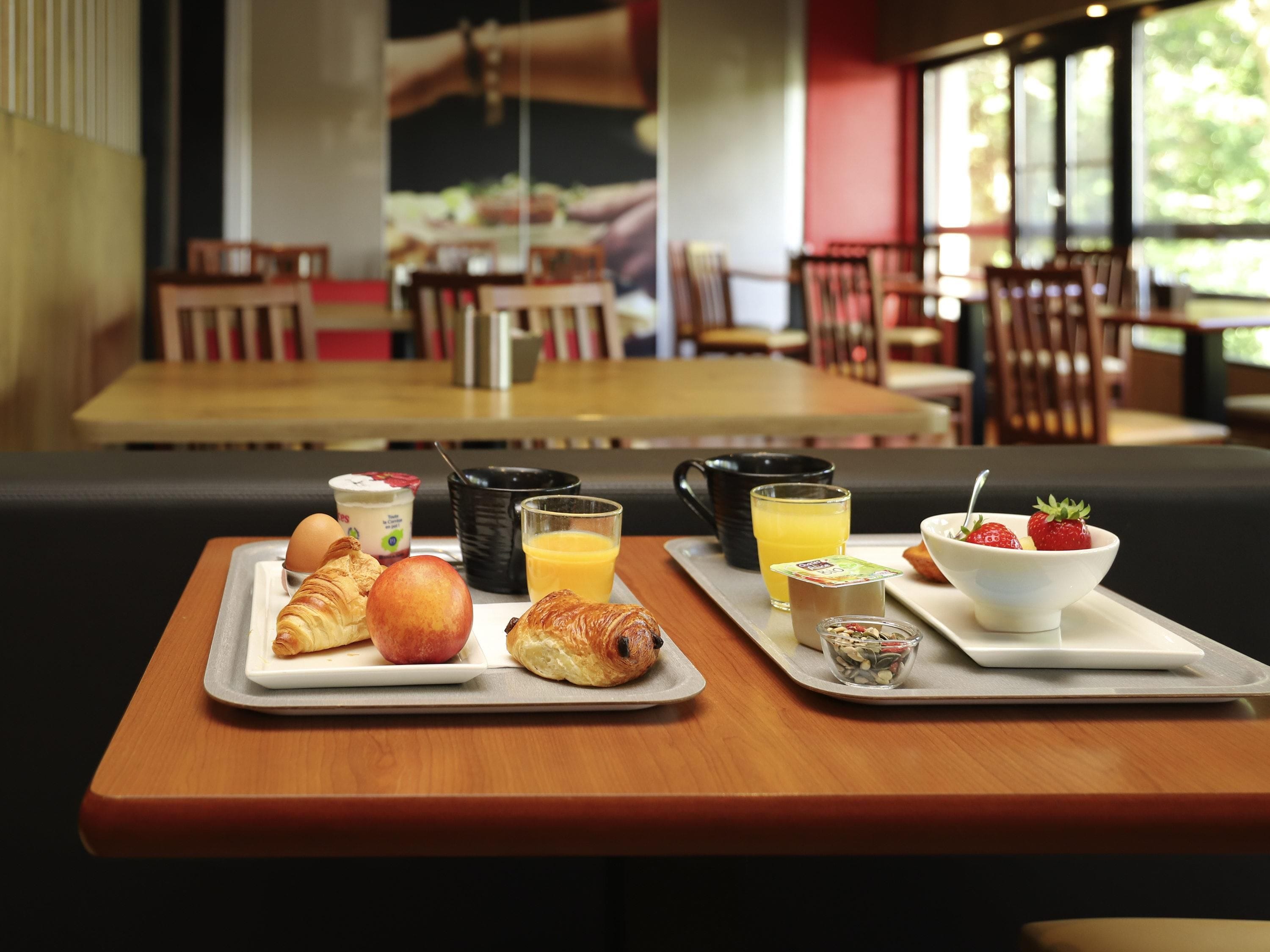 A breakfast tray with croissants, an apple, juice, yogurt, and a bowl of strawberries on a wooden table in a restaurant.