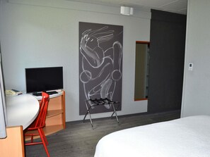 A hotel room with a bed, a desk with a TV, a chair, a wall-mounted artwork, and a door.