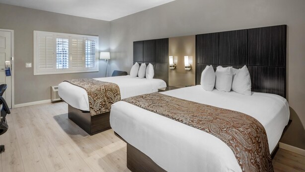 Habitación de hotel con dos camas, un ventanal con persianas, un televisor de pared y una mesita de noche.