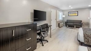 Una habitación de hotel moderna con un aparador de madera oscura, un televisor de pantalla plana, un sofá gris, una cama con ropa de cama estampada y una ventana con persianas.
