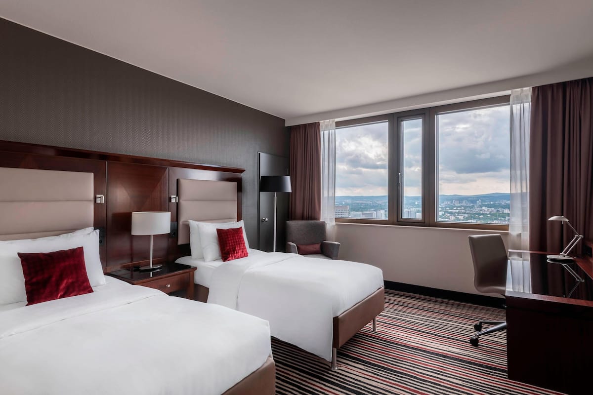 Een hotelkamer met twee bedden, een bureau, een stoel en een groot raam met uitzicht op de stad.