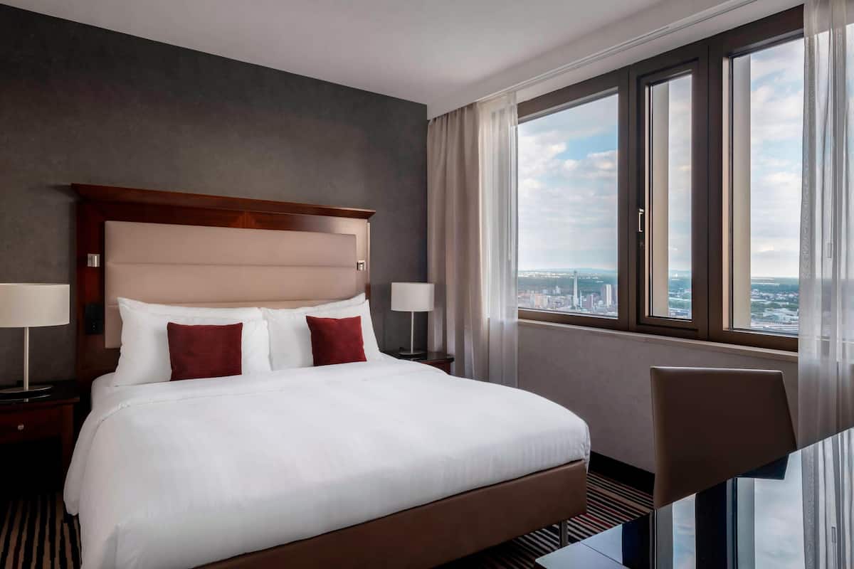 Hotelkamer met een groot bed, twee nachtkastjes met lampen, een stoel en een groot raam met uitzicht op de stad.