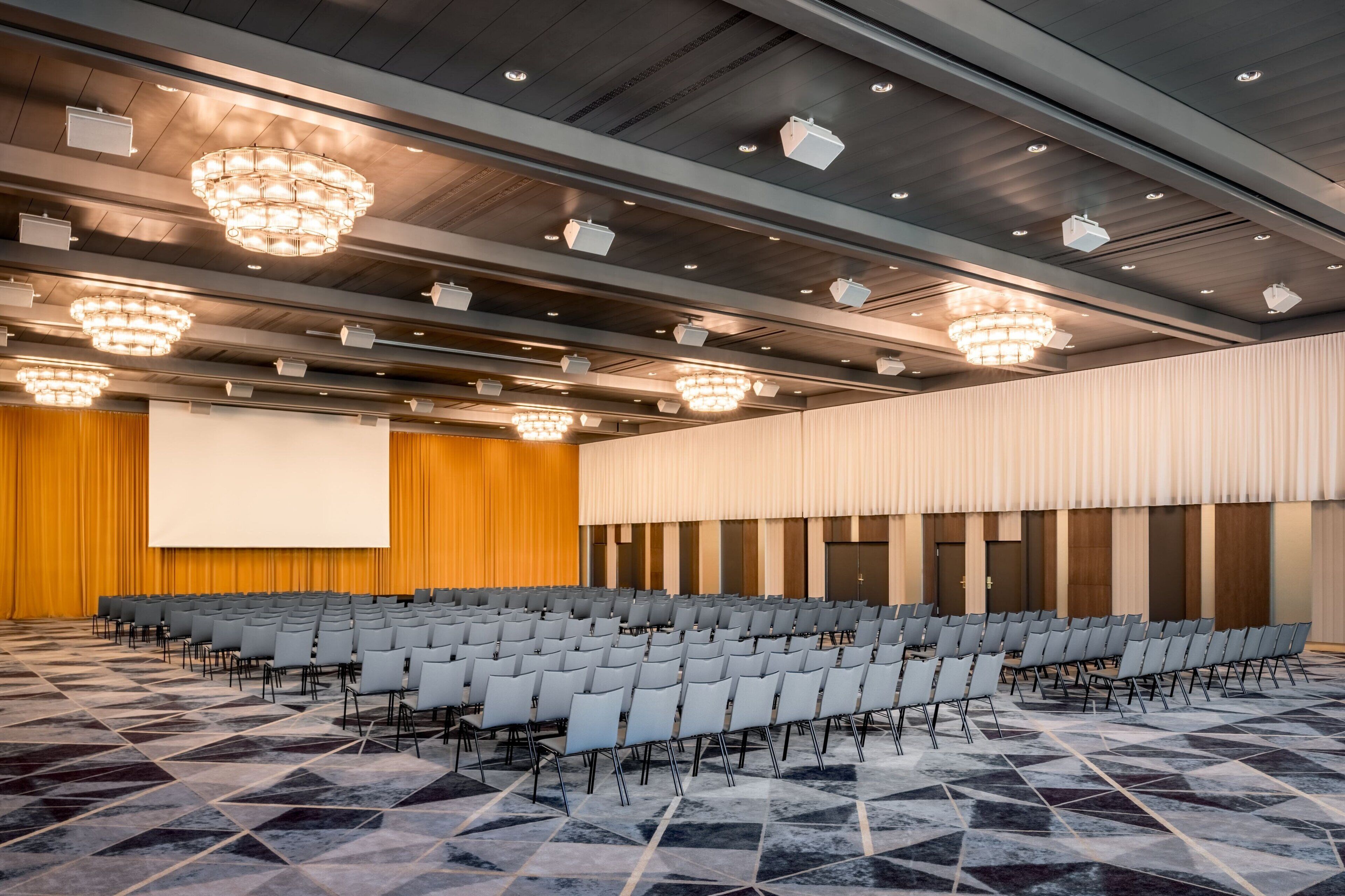 Uma sala de conferências com fileiras de cadeiras, uma tela grande e lustres.