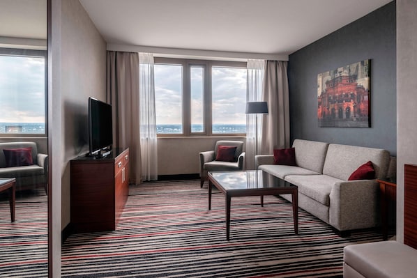 Habitación de hotel con un ventanal grande, un televisor de pantalla plana, un sofá, una silla, una mesita de centro y una alfombra estampada.