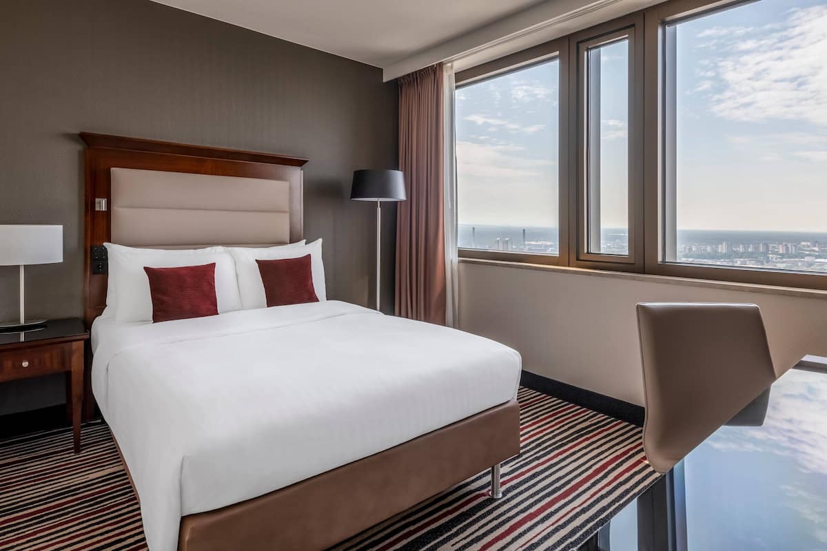 Een hotelkamer met een groot bed, een bureau, een stoel en uitzicht op de stad door grote ramen.