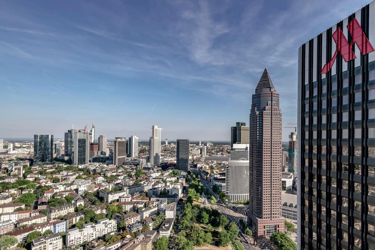 Stadsgezicht met moderne wolkenkrabbers, waaronder een opvallende toren met een puntige top en een groot rood logo op een gebouw aan de rechterkant.