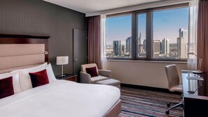Ein Hotelzimmer mit einem großen Bett, zwei Sesseln, einem Schreibtisch mit Stuhl, einer Lampe und Blick auf eine Stadtansicht durch ein großes Fenster.
