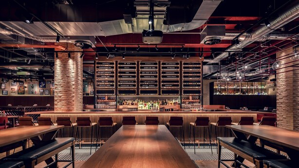 Eine Bar mit einem langen Holztisch und mehreren Barhocker, umgeben von Regalen voller Flaschen.