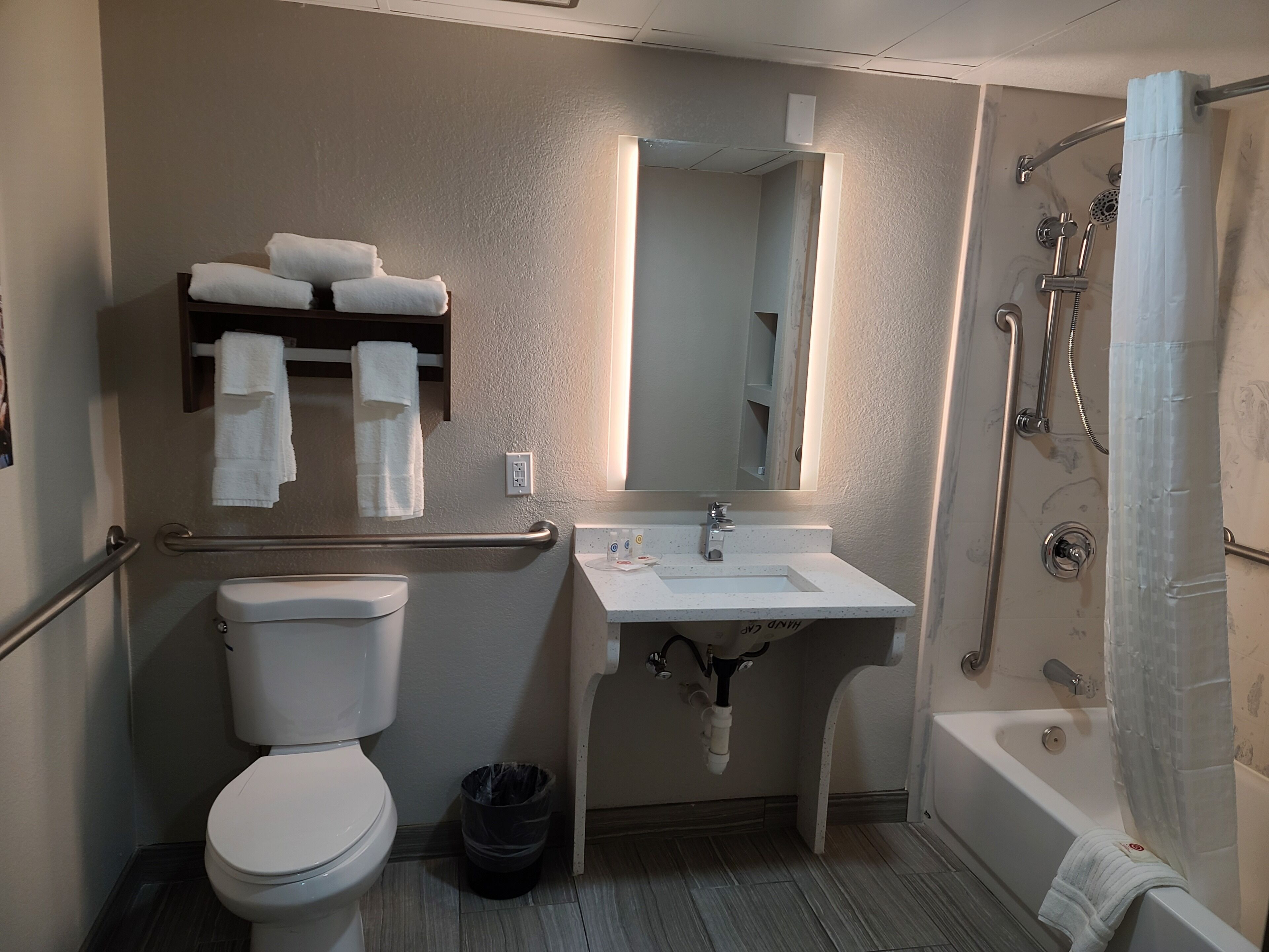 Een badkamer met toilet, wastafel en douche. Handdoeken liggen op een plank en er staat een prullenbak.