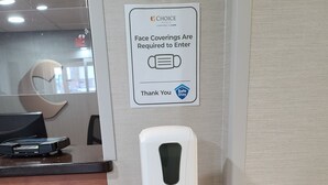 A sign indicating face coverings are required to enter, a hand sanitizer dispenser, and a reception desk with a laptop.