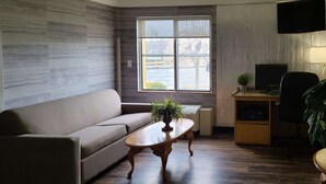 A modern living room with a grey sofa, a wooden coffee table with a plant, a desk with a computer, and a large window with a view of the outdoors.
