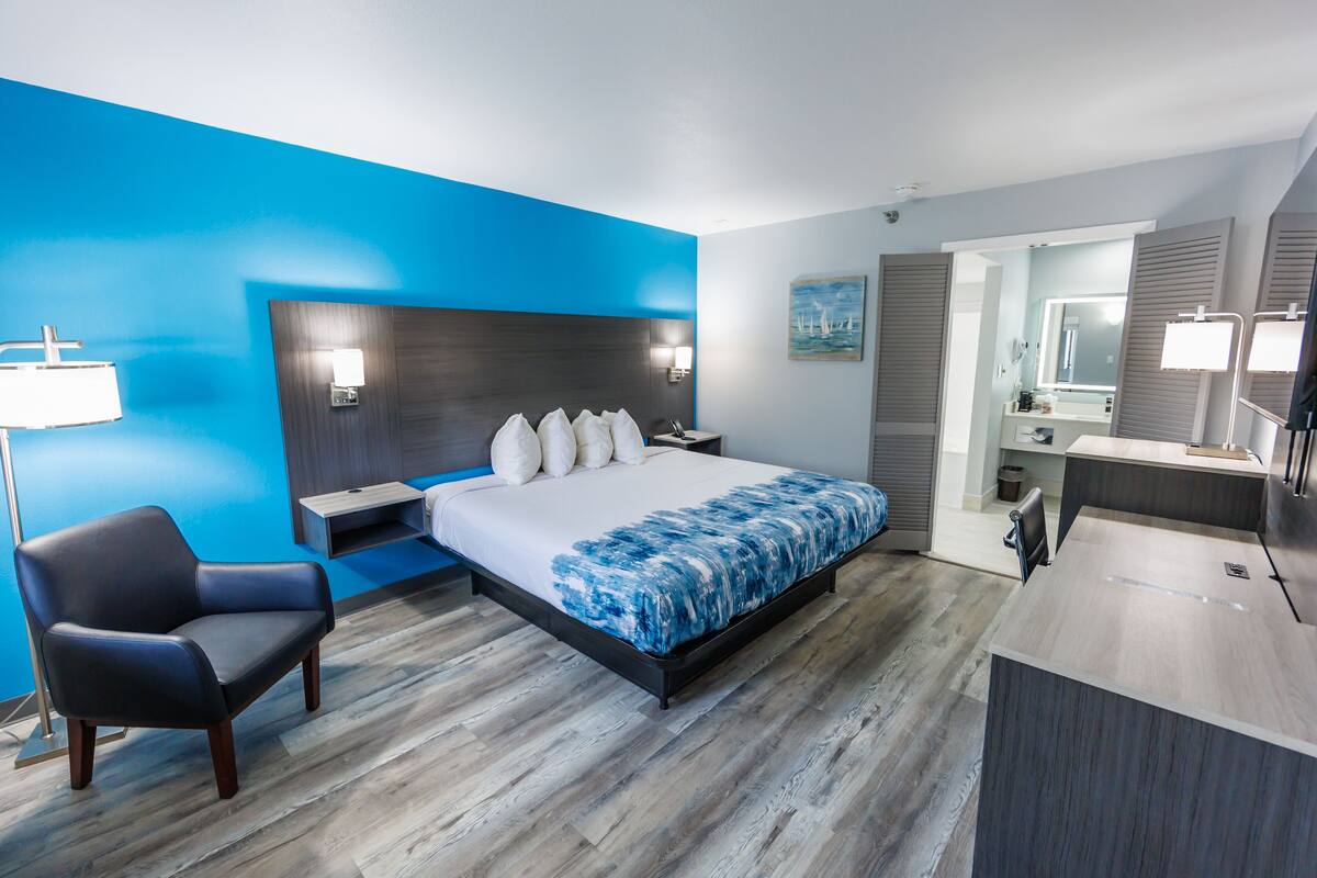 Habitación de hotel moderna con cama, escritorio, silla, mesita de noche y baño visible a través de una puerta abierta.