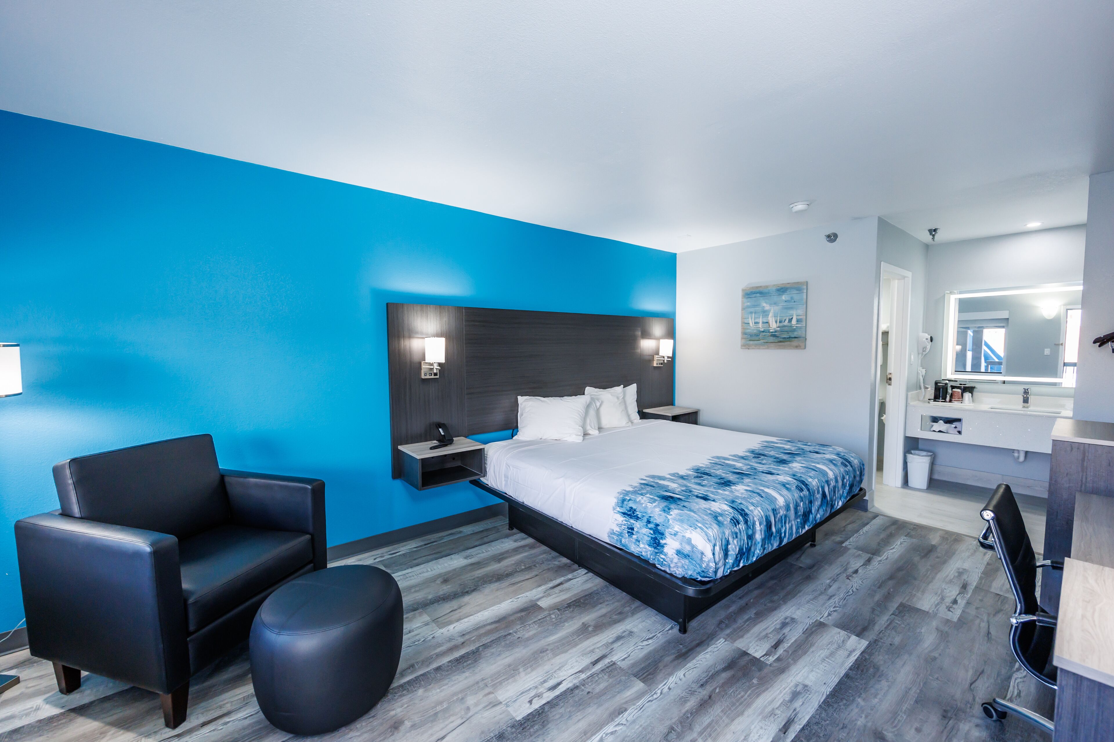 A modern hotel room with a large bed, a black leather sofa, a small ottoman, and a bathroom with a mirror and sink.