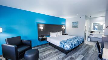 A modern hotel room with a large bed, a black leather sofa, a small ottoman, and a bathroom with a mirror and sink.