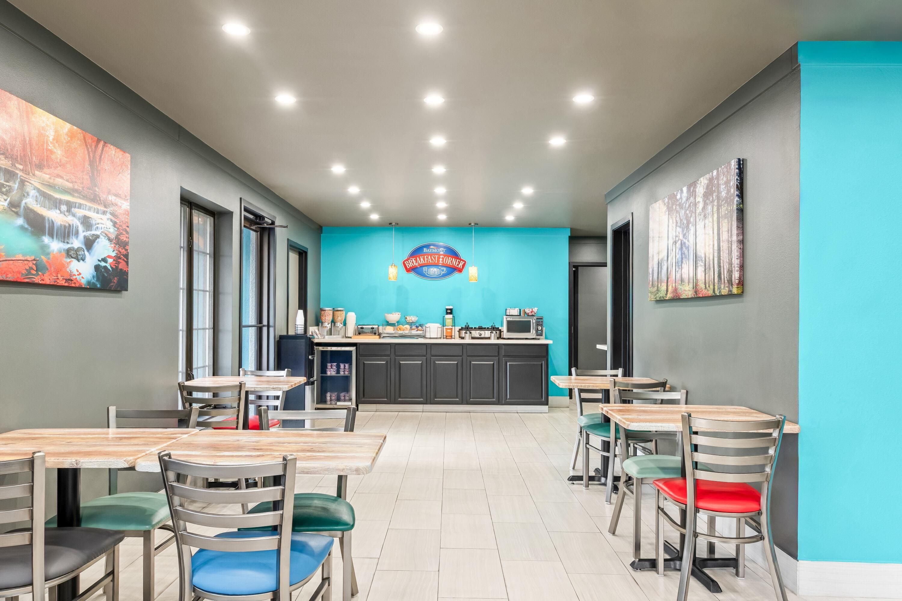 Cafetería para el desayuno con mesas y sillas, un área de cocina y un letrero que dice 'Baymont Breakfast Cafe'.