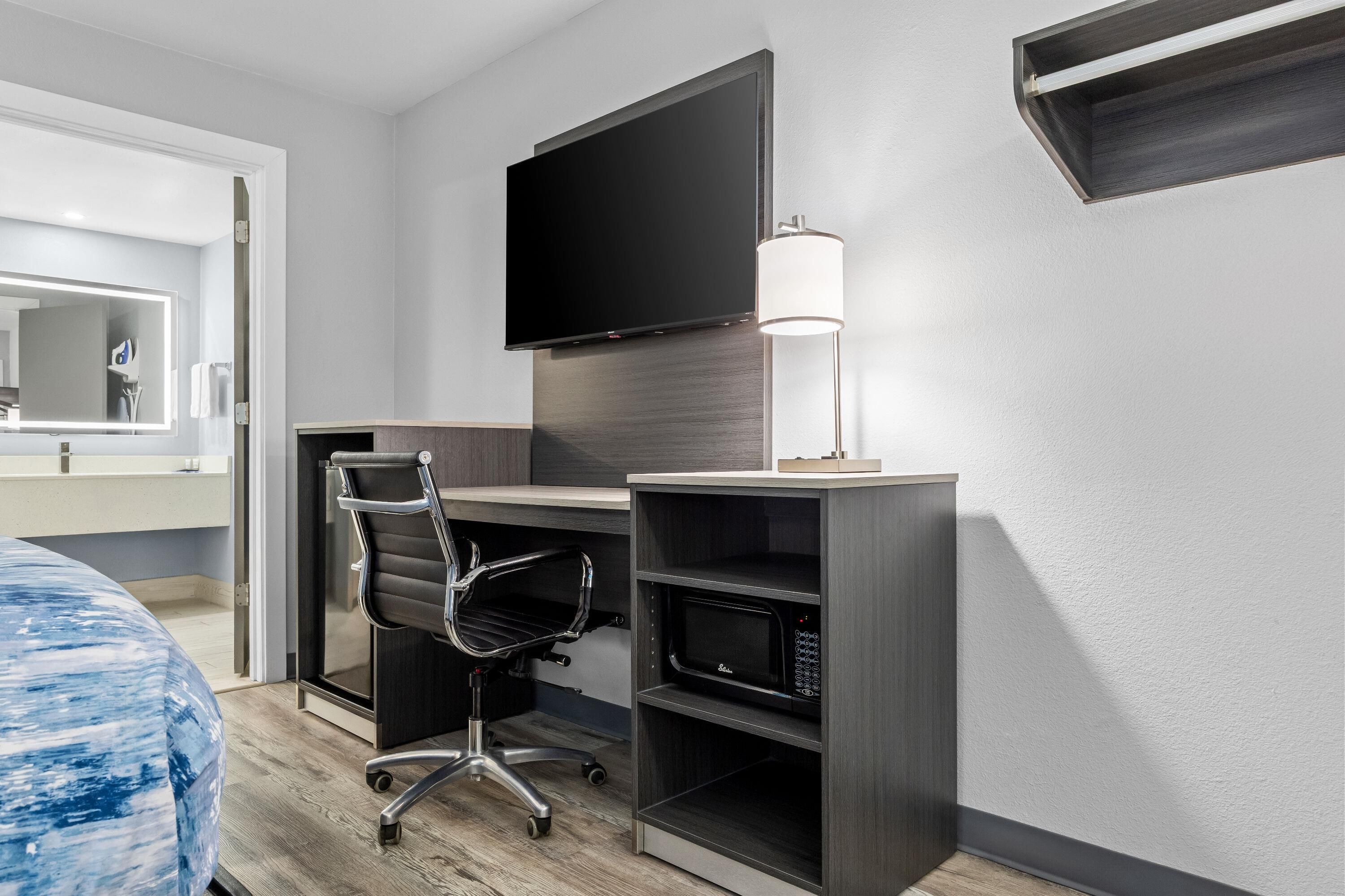 A hotel room with a desk, chair, flat-screen TV, and a lamp.