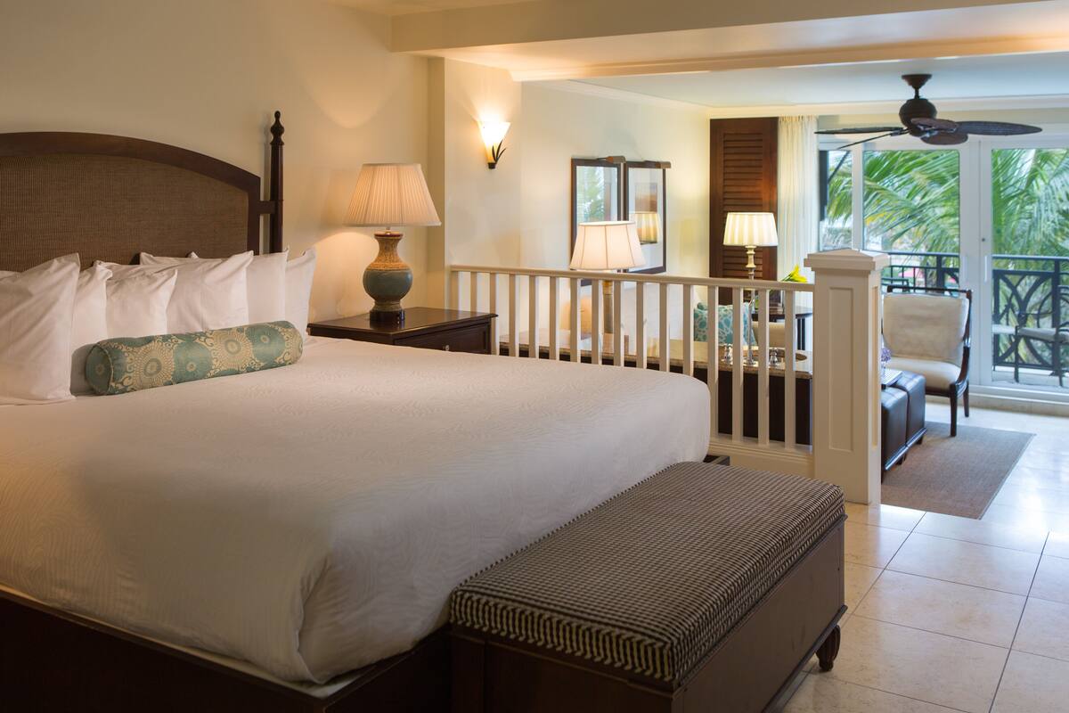 A hotel room with a large bed, a bench, a balcony with a chair, and a view of palm trees.