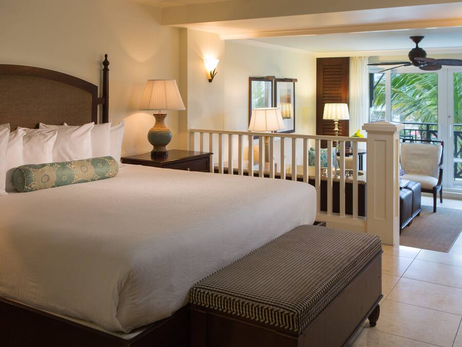 Habitación de hotel con una cama grande, una banca, un balcón con una silla y vistas a palmeras.